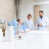 Medikamentenflaschen und Messbecher mit Tabletten auf Tisch im Krankenhauszimmer – medizinische Versorgung mit Arzt und Patientin im Hintergrund.