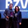 Zwei Personen auf Bühne mit Auszeichnung vor großem Logo der RZV Rechenzentrum Volmarstein GmbH, beide in dunkler Kleidung mit Namensschildern.