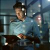 Mann in hellblauem Hemd arbeitet nachts mit Tablet in modernem Bürogebäude – reflektierende Glaswand und Lichter im Hintergrund vermitteln geschäftige Arbeitsatmosphäre