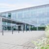 Modernes Bürogebäude mit Glasfassade und überdachtem Eingangsbereich, umgeben von gepflastertem Vorplatz und grüner Vegetation im Vordergrund.