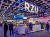 Messestand von RZV auf einer großen Messehalle mit modernem Design und auffälliger Beleuchtung. Über dem Stand hängt ein leuchtender Rahmen mit dem Schriftzug ‚RZV‘. Im Bereich sind mehrere Infotheken, Sitzgelegenheiten und Pflanzen zu sehen. Zahlreiche Besucher stehen und unterhalten sich. Im Hintergrund sind weitere Messestände und Banner mit Themen wie ‚Digitalisierung im Gesundheitswesen‘ sichtbar.