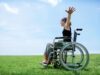 Person im Rollstuhl auf einer grünen Wiese unter blauem Himmel mit ausgestrecktem Arm – Symbol für Freiheit, Inklusion und Barrierefreiheit.