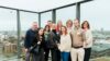 Gruppe von Personen steht auf einer Dachterrasse mit Glasgeländer und Blick über eine Stadtlandschaft. Im Hintergrund sind Gebäude, Straßen und ein Fernsehturm zu sehen. Die Personen tragen überwiegend Freizeit- oder Business-Casual-Kleidung und stehen eng zusammen für ein Gruppenfoto.