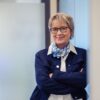 Eine Person in geschäftlicher Kleidung steht mit verschränkten Armen in einem Büroflur. Sie trägt eine dunkle Jacke über einer hellen Bluse und ein gemustertes Halstuch.