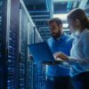 Zwei Personen arbeiten mit einem Laptop in einem Serverraum vor mehreren beleuchteten Serverracks.