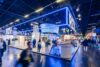 Großer Messestand mit blau beleuchtetem Rahmen und dem Schriftzug „RZV“ in einer modernen Ausstellungshalle. Zahlreiche Besucherinnen und Besucher bewegen sich zwischen Theken, Sitzbereichen und Pflanzenarrangements, während im Hintergrund weitere Ausstellerstände und Beleuchtungselemente zu sehen sind.