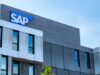 Außenansicht eines modernen SAP-Firmengebäudes mit markantem SAP-Logo auf der oberen Gebäudeecke. Die Fassade ist in Grau- und Silbertönen gestaltet und wird von großen Fenstern geprägt. Im Vordergrund ist ein kleiner Bereich einer Baumkrone sichtbar, darüber erstreckt sich ein klarer blauer Himmel.