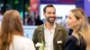 Drei Personen stehen auf einer Fachmesse in einem Gespräch zusammen. Im Fokus steht eine Person mit Namensschild, während Messestände und weitere Besuchende im Hintergrund unscharf zu sehen sind.