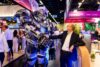 Eine Person in Business-Kleidung steht auf einer Messe und schüttelt einem großen futuristischen Roboter die Hand. Im Hintergrund sind Messestände, Besuchende und beleuchtete Ausstellungsflächen zu sehen.