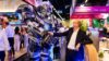 Eine Person in Business-Kleidung steht auf einer Messe und schüttelt einem großen futuristischen Roboter die Hand. Im Hintergrund sind Messestände, Besuchende und beleuchtete Ausstellungsflächen zu sehen.
