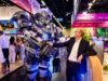Eine Person in Business-Kleidung steht auf einer Messe und schüttelt einem großen futuristischen Roboter die Hand. Im Hintergrund sind Messestände, Besuchende und beleuchtete Ausstellungsflächen zu sehen.